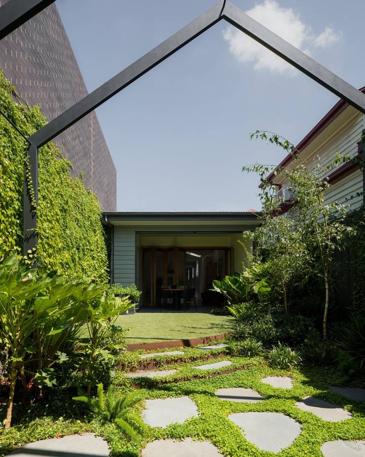 A small back garden with stone steppers set in green groundcover, a lawn, large leaved plants and a wall of climbers, under a dark steel gable frame, looking to a weatherboard house with folding glass doors open to a dining room.