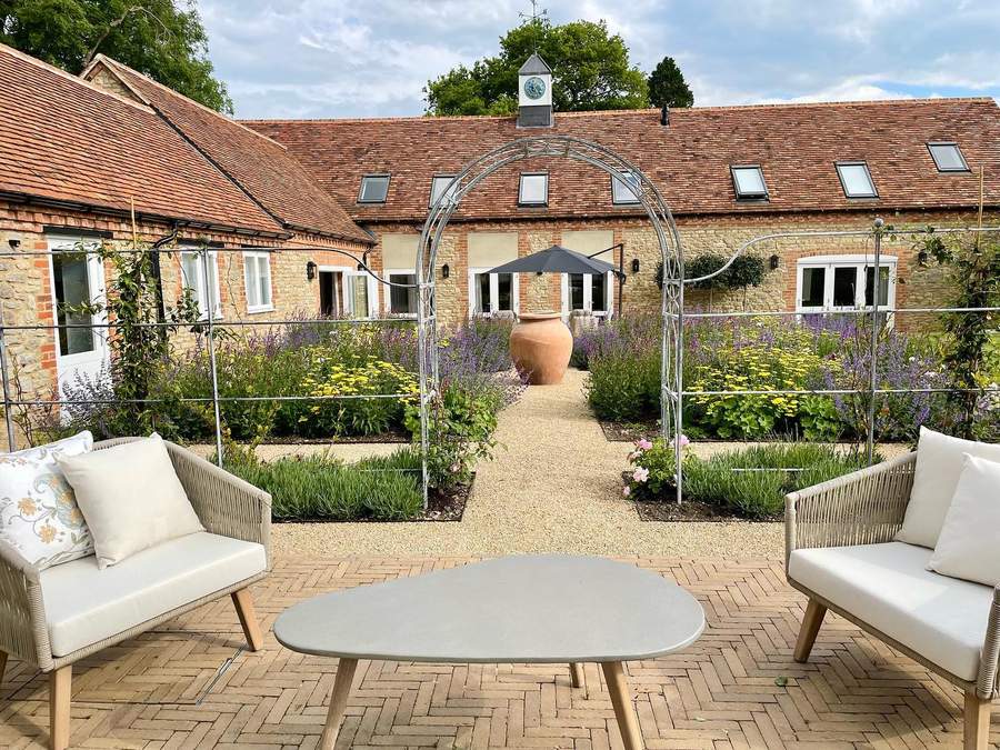 A brick paved terrace with two woven outdoor sofas and an oval table, looking along a gravel path under a metal arch to a terracotta urn, with beds of lavender, yellow green alchemilla and roses, and a stone barn with a tiled roof and a clock turret behind.