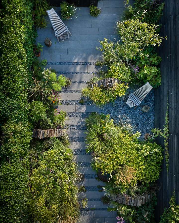 An overhead view of a long narrow garden with a path of dark concrete slabs and gravel bands running down the middle, beds of grasses, shrubs and flowering perennials either side edged with low logs, two pale timber loungers, a climber covered boundary on one side and a dark timber fence on the other.