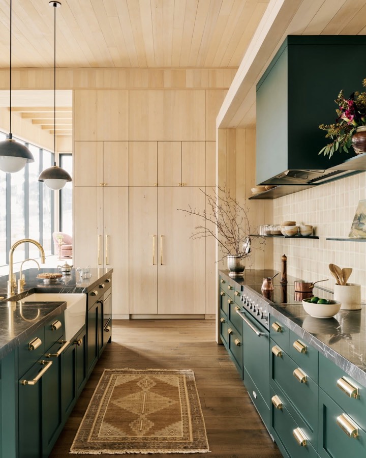 A kitchen with deep teal cabinets and range, pale timber joinery above and a patterned rug on the floor.