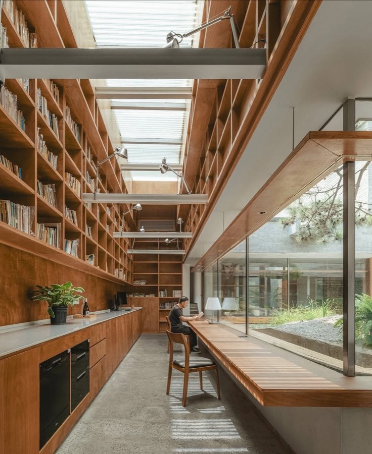 A long room lined with timber bookshelves under skylights, opening through full height glass to a courtyard.