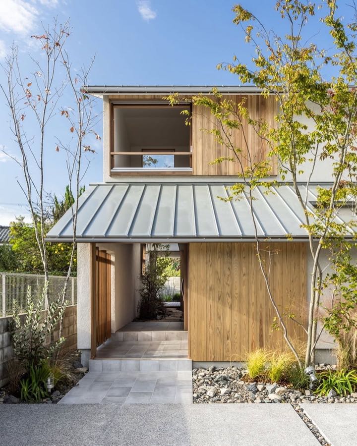 A timber clad house with a low metal awning over the entry path, with young trees beside it.
