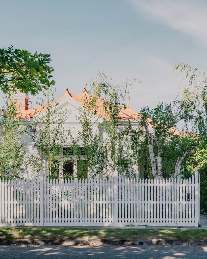 A period house behind a white picket fence and young trees, with a tiled roof above the greenery.