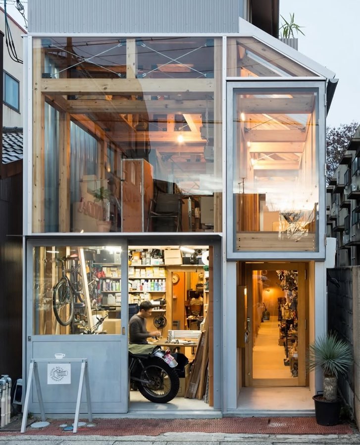 A narrow two-storey glass and steel shopfront lit from inside at dusk, with a motorbike in the workshop.