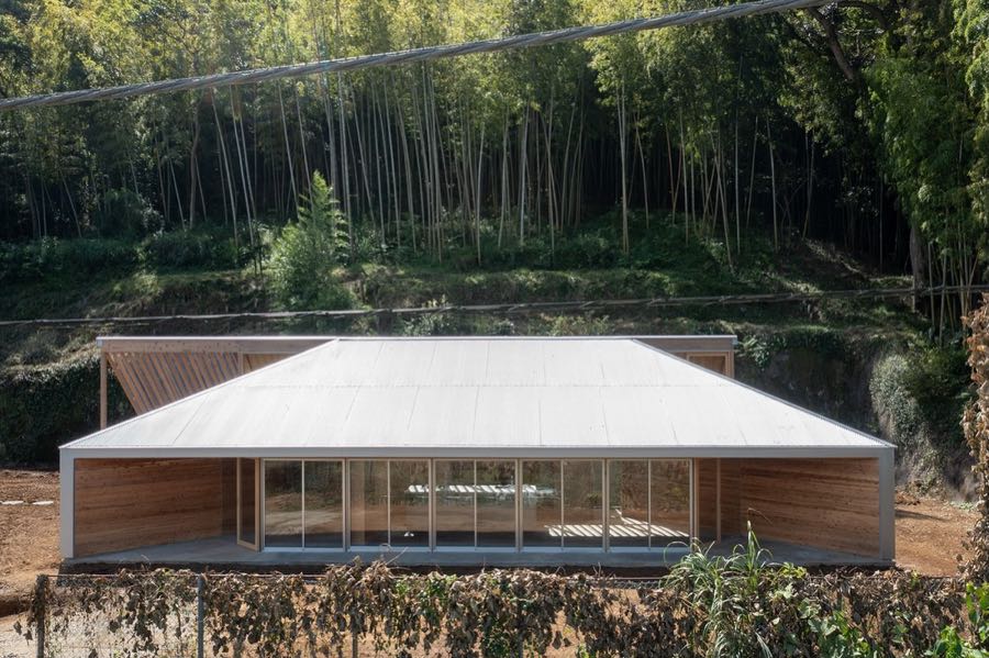A low building with a wide shallow gable roof set on a terrace below a bamboo hillside.