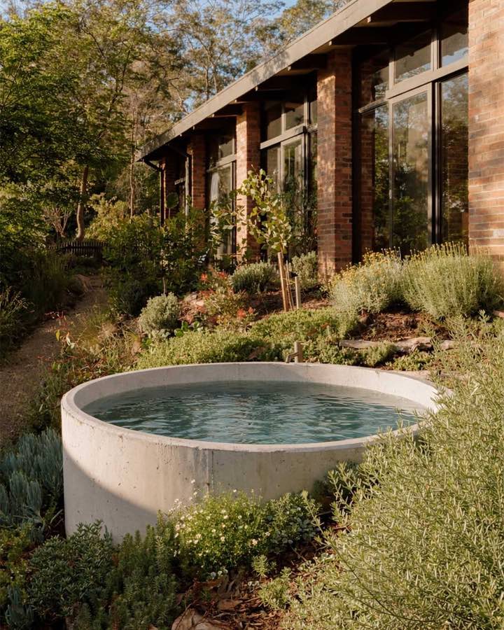 A round concrete outdoor bath set into a planted garden beside a long brick and timber house.