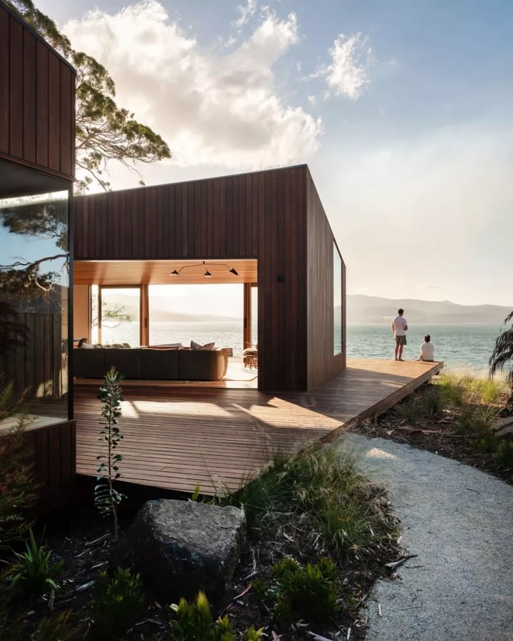 A dark timber holiday house on a deck above a beach, with a person standing at the edge looking out.