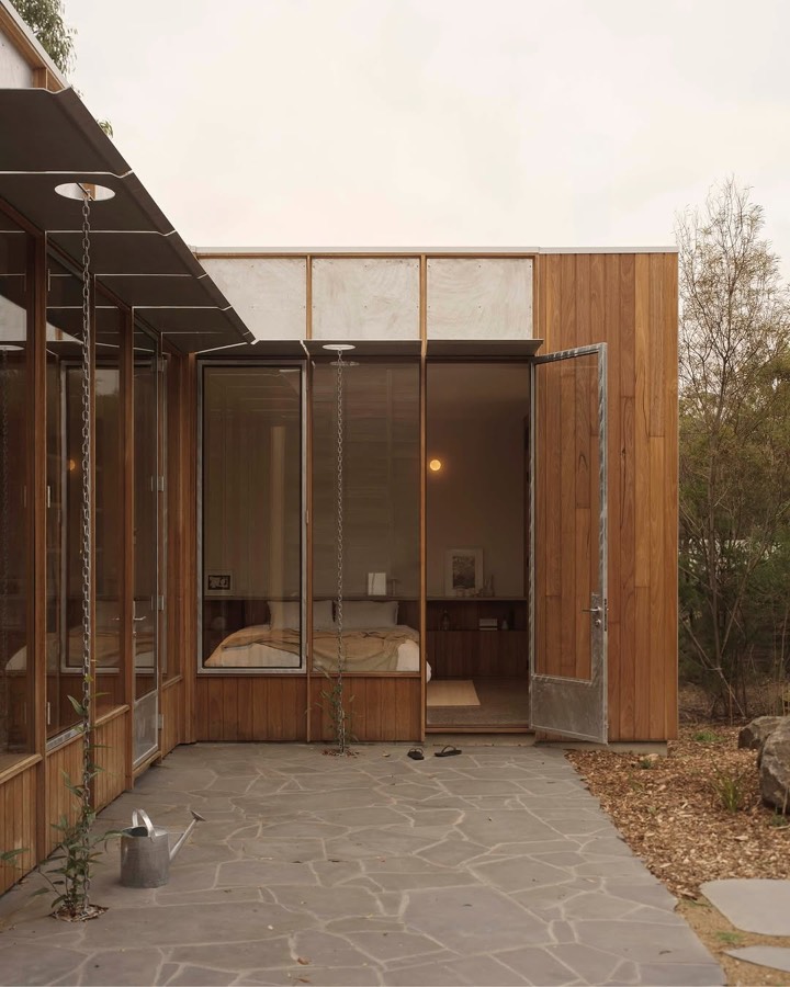 A small timber and glass cabin with doors folded open onto crazy paving, with a bed visible inside.