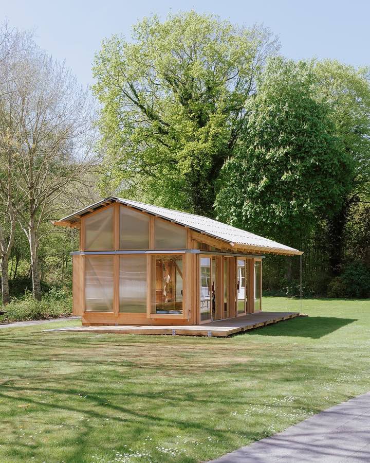 A small timber pavilion with a pitched roof standing on mown grass in front of tall trees.