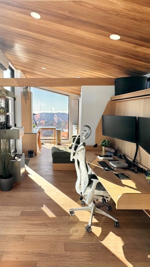 A timber lined interior with a two-monitor desk and an office chair, looking out to mountains.