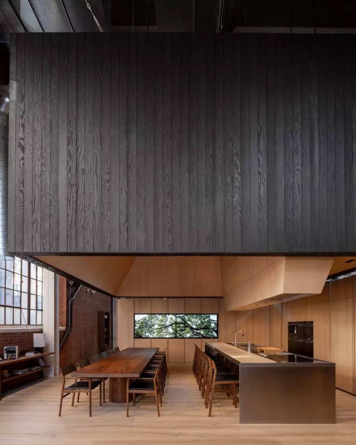 A dark timber box hanging above a long dining table and a pale kitchen bench in a converted industrial building.