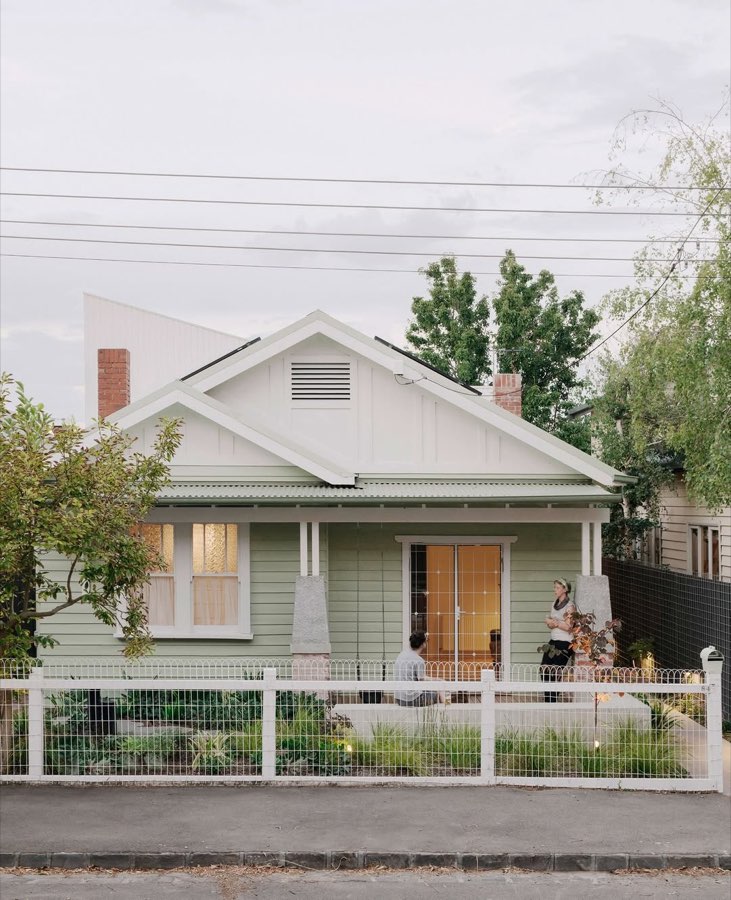A pale green weatherboard cottage behind a white post and wire fence, with people on the verandah.