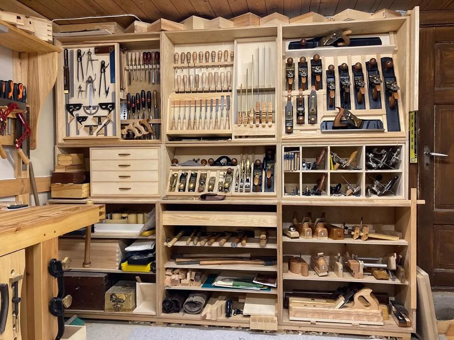A large timber tool wall holding chisels, planes and saws in fitted racks, floor to ceiling beside a workbench.
