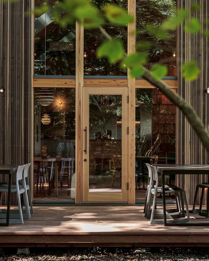 A timber and glass building lit from within, with outdoor tables and chairs on a deck.