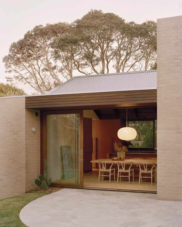 A pale brick house with a sliding glass wall open to a dining table under a round paper pendant, with moonah trees behind.