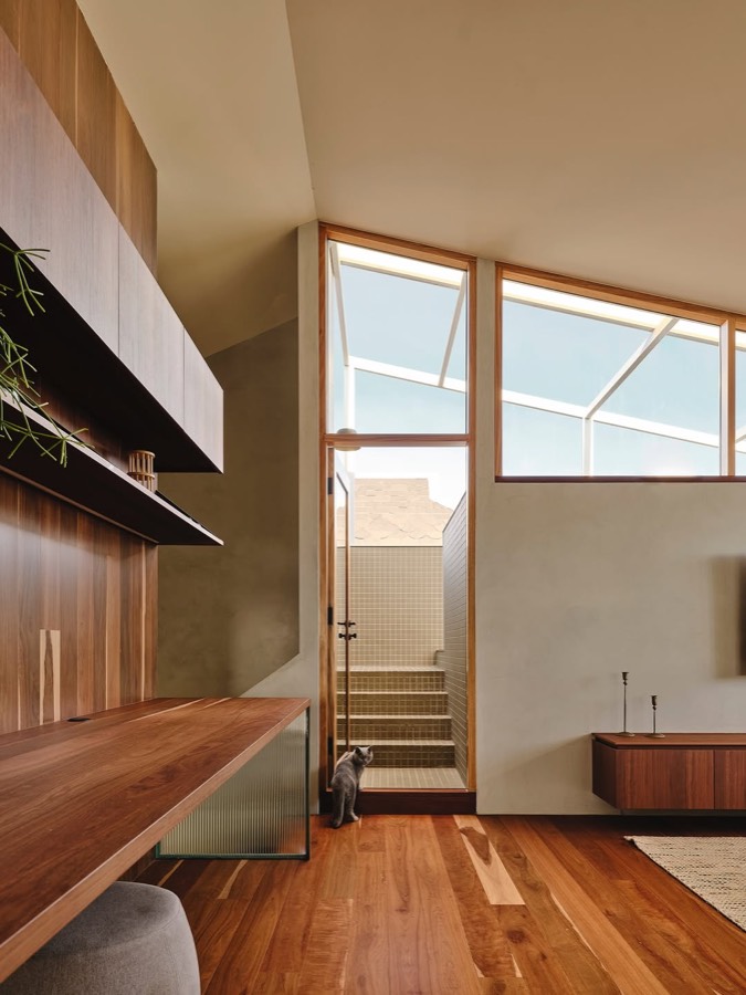 A timber lined room with a long desk and shelves, looking through to a stair under a clerestory window.