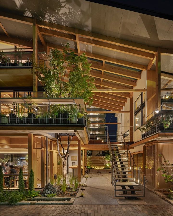 A timber framed building of stacked open decks planted with greenery, lit at night, with a steel stair between.