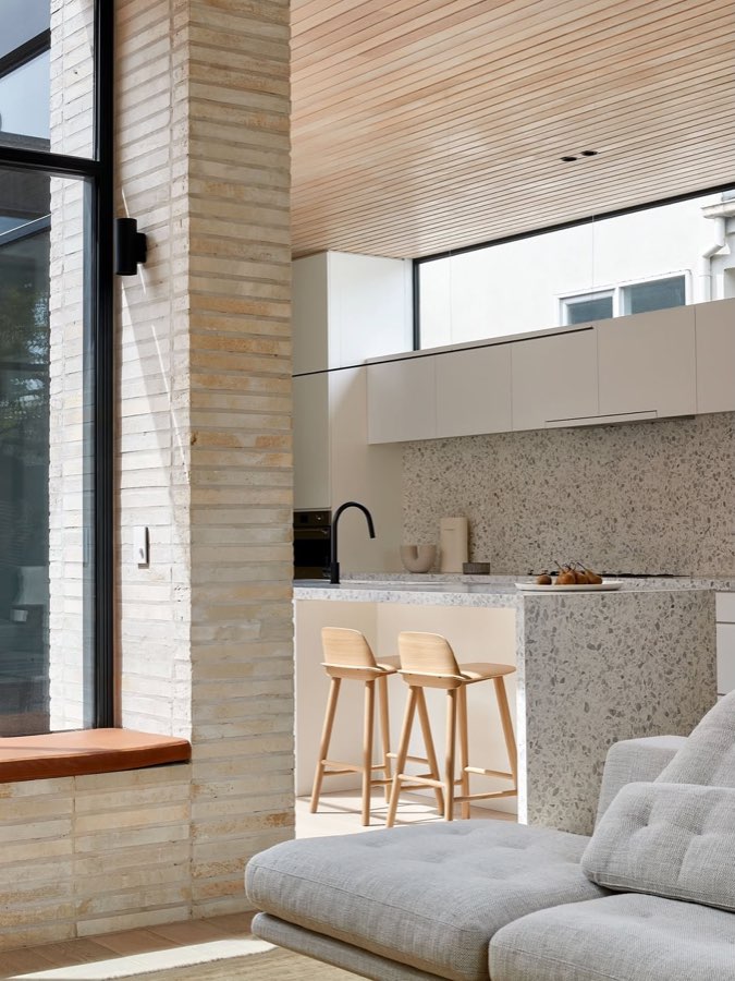A kitchen in terrazzo and pale brick with timber stools, seen past a brick pier from a living room.