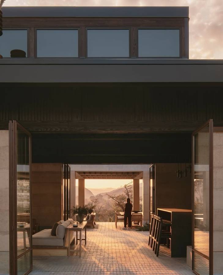 A house at dusk with folding glass doors open onto a paved living space, looking through to distant hills.