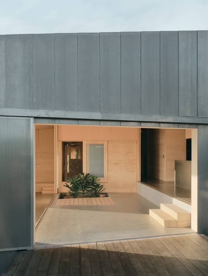 A grey clad house opening to a plywood lined courtyard room with a planting bed and a timber deck.