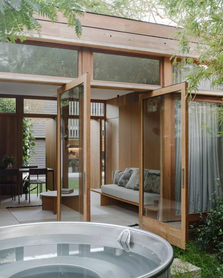 A timber and glass pavilion with pivoting doors open to a daybed inside, and a round plunge tub outside.