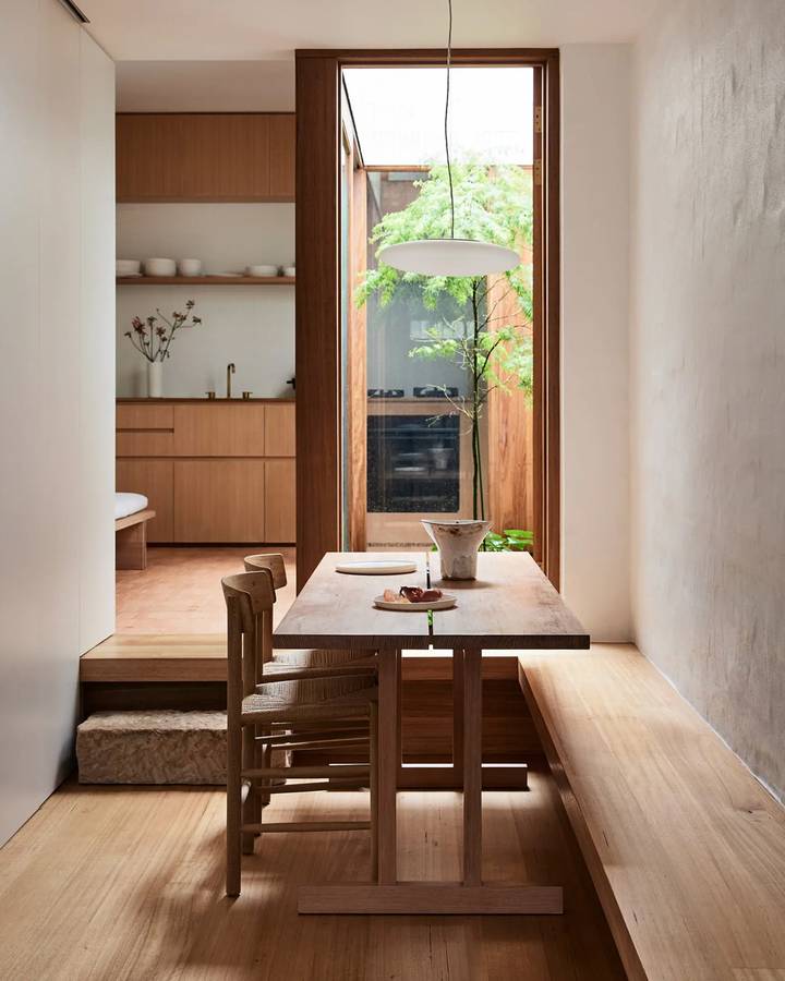 A narrow dining room with a timber table and bench, a tall window to a courtyard and a kitchen beyond.