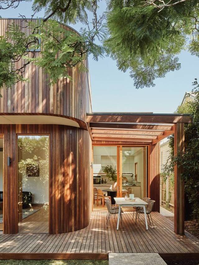 A curved timber clad upper floor above a pergola and deck, with a kitchen through open glass doors.