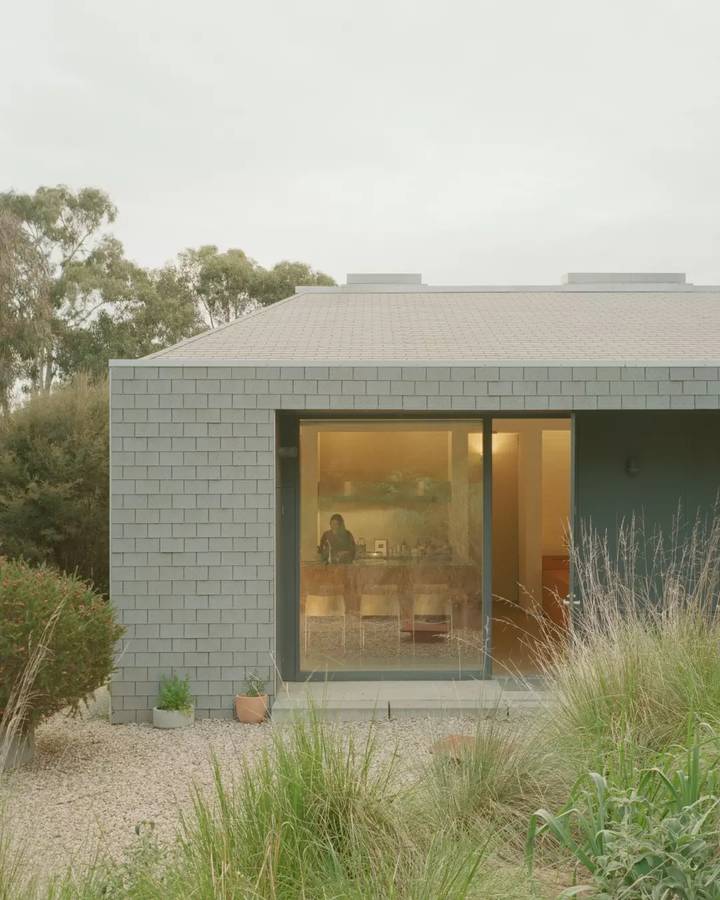 A low house clad in grey shingles, with a wide sliding window onto a kitchen, standing behind grasses and gravel.