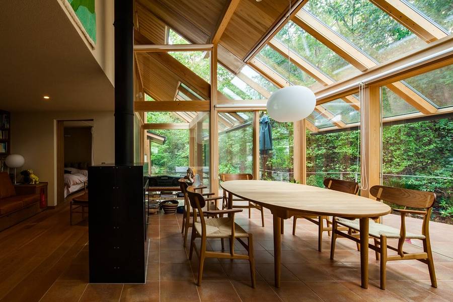 A dining room under a glazed timber roof, with an oval table, woven chairs and woodland outside.