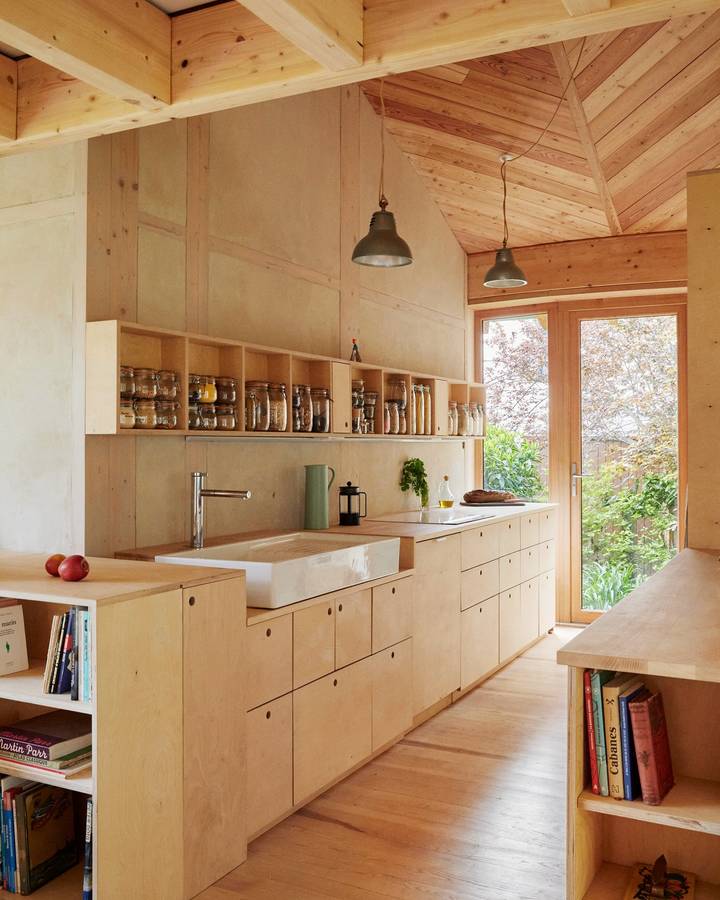 A plywood lined kitchen with a long bench, a shelf of jars and a glazed door to the garden.