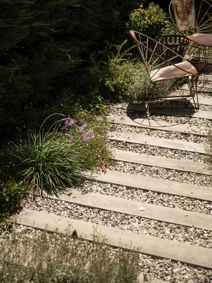 A path of concrete planks set in gravel through low planting, with a rusted wire chair beside it.