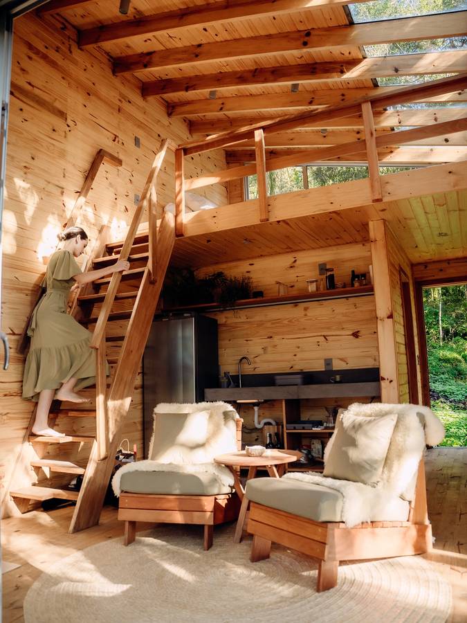 A pine lined cabin interior with a ladder stair to a loft, two timber armchairs and a small kitchen.