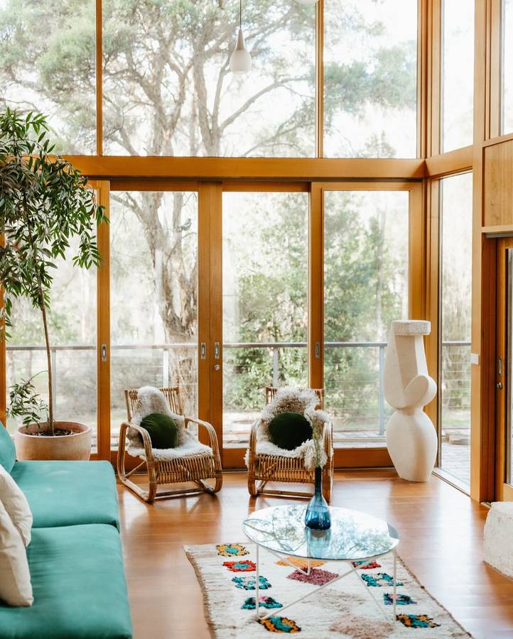 A living room with full height timber framed glazing to bushland, cane armchairs and a green sofa.